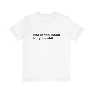 Puede incluir: Una camiseta blanca de manga corta con el texto en negro "Not in the mood for your shit." impreso en una fuente sans-serif sencilla en el centro del pecho. La prenda tiene un cuello redondo y una silueta de corte recto estándar.