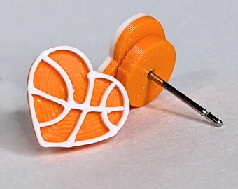 May include: Orange and white heart-shaped stud earrings with a basketball design. The earrings feature a basketball graphic in white lines on an orange heart shape. The post is silver-colored.