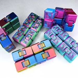 May include: A collection of five multi-colored infinity cube fidget toys. Each toy is composed of several interconnected plastic blocks featuring a gradient of vibrant hues including pink, blue, green, and purple with a metallic, iridescent finish.