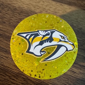 Può includere: Un'impugnatura per telefono rotonda, di colore giallo glitterato, con un logo dei Nashville Predators in bianco e nero.
