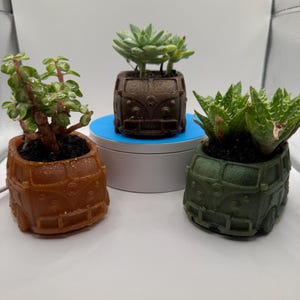May include: Three small potted plants in the shape of vintage Volkswagen vans. The vans are brown, green, and orange. The plants are succulents.
