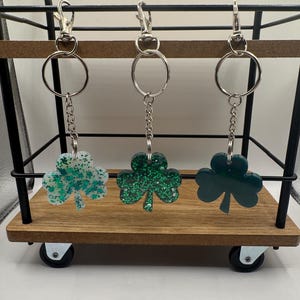 Może przedstawiać: Trzy breloki w kształcie koniczyny z zielonym brokatem, jeden z białym brokatem, wiszące na srebrnych kółkach do kluczy na czarnej metalowej półce.