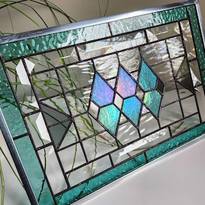 May include: Stained glass window with a geometric design. The window features a silver metal frame and a pattern of clear, teal, and iridescent glass pieces. The central design includes diamond and triangular shapes.