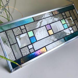May include: Rectangular stained glass panel with a silver metal frame. The design features a geometric pattern with clear, textured, and iridescent glass in shades of blue, green, purple, and silver. A chain is attached for hanging.