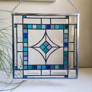 May include: Square stained glass panel with a geometric design. The panel features a central diamond shape and is framed with a silver metal border. The design incorporates shades of blue, teal, and white glass, creating a decorative piece.