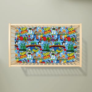 May include: A light blue cot sheet featuring a repeating pattern of Toy Story Mania characters including Woody, Buzz Lightyear, and Slinky Dog, alongside colourful text logos, stars, and balloons, all contained within a light-coloured wooden cot frame.