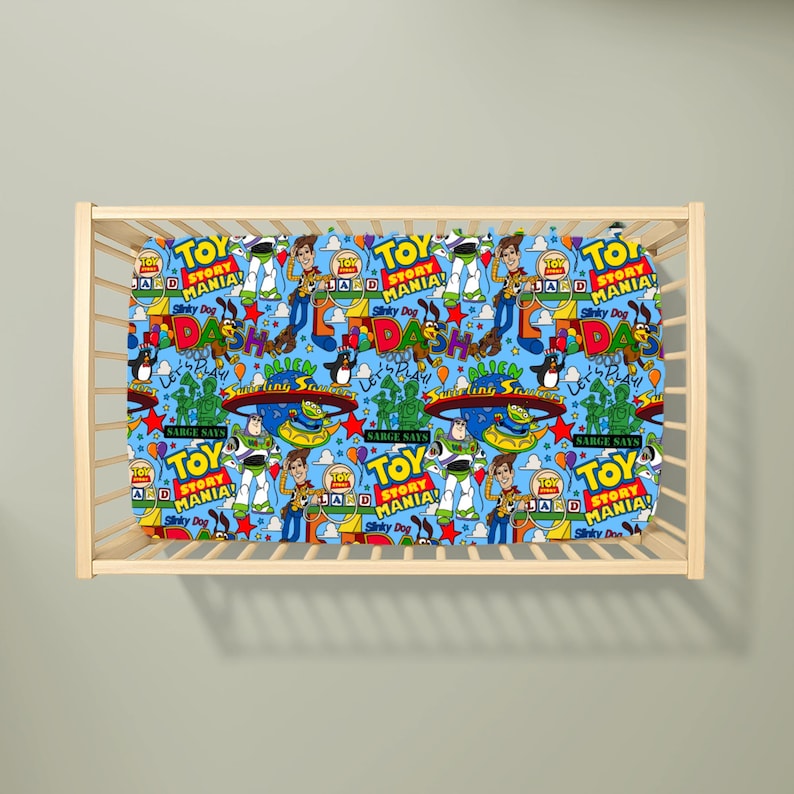 May include: A light blue cot sheet featuring a repeating pattern of Toy Story Mania characters including Woody, Buzz Lightyear, and Slinky Dog, alongside colourful text logos, stars, and balloons, all contained within a light-coloured wooden cot frame.