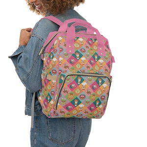 May include: A pink and patterned backpack featuring a colorful diamond pattern with various cartoon character faces. The backpack has pink straps and a top handle, with a front pocket and side zippers. The bag is worn over the shoulders.