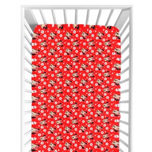 May include: A red fitted crib sheet with a repeating pattern of Minnie Mouse faces and white flower shapes. The sheet is inside a white crib frame, showcasing the design's coverage and scale. The overall theme is children's bedding.
