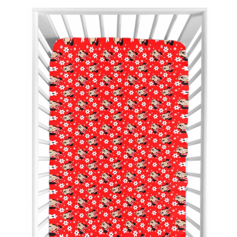 May include: A red fitted crib sheet with a repeating pattern of Minnie Mouse faces and white flower shapes. The sheet is inside a white crib frame, showcasing the design's coverage and scale. The overall theme is children's bedding.