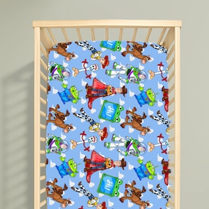 May include: A light blue cot sheet featuring a repeating pattern of cartoon characters including a space ranger, a cowboy, a cowgirl, a green alien, a white spork character, a brown horse, and a green square sign with the text Hi printed on it.