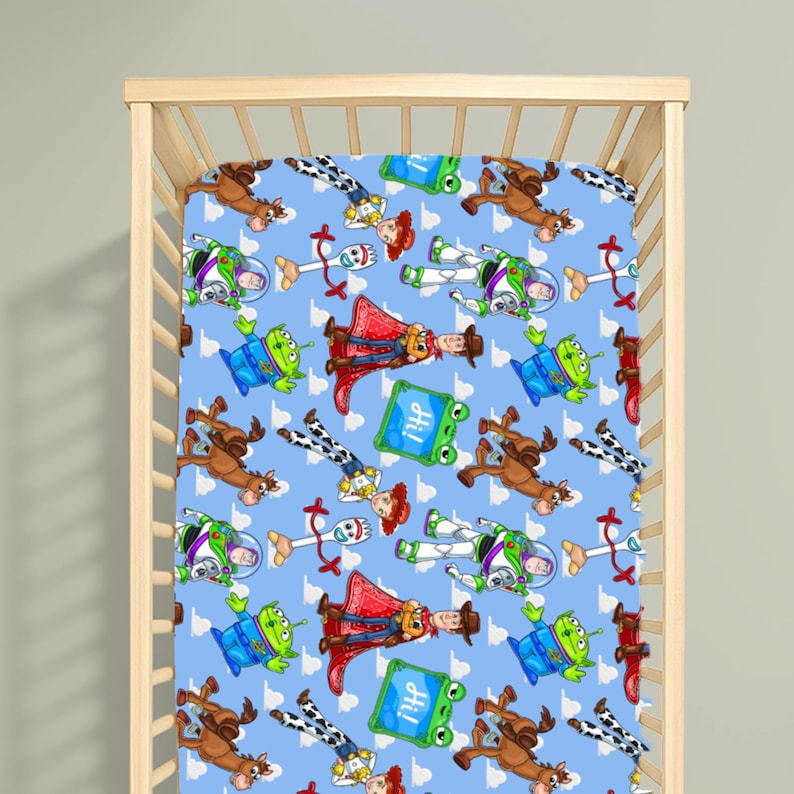 May include: A light blue cot sheet featuring a repeating pattern of cartoon characters including a space ranger, a cowboy, a cowgirl, a green alien, a white spork character, a brown horse, and a green square sign with the text Hi printed on it.