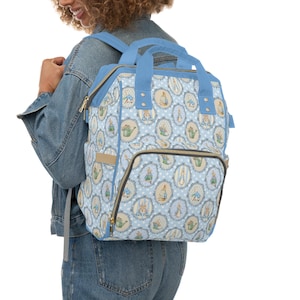 May include: A light blue backpack with a repeating pattern of oval illustrations featuring a rabbit, watering can, and other garden elements. The bag has blue straps, handles, and trim, with a beige pocket and gold zippers.