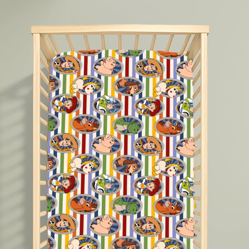 Puede incluir: Una s&aacute;bana de cuna con un patr&oacute;n repetitivo de personajes de Toy Story, incluyendo a Woody, Buzz Lightyear, Rex, Hamm, Perdig&oacute;n y Jessie, cada uno en un marco ovalado. El fondo presenta rayas verticales en colores rojo, amarillo y verde.