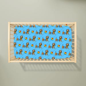May include: A light blue cot sheet featuring a repeating pattern of brown dachshunds with spring-like bodies and colourful balls with red stars. The design is set against a background of thin, dark blue dashed lines forming a diamond grid pattern.