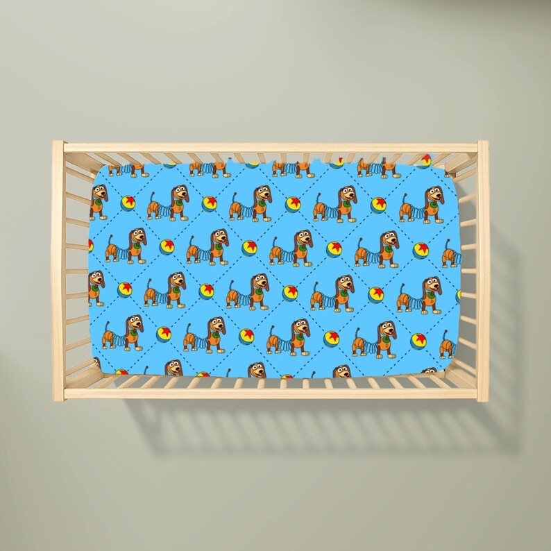 May include: A light blue cot sheet featuring a repeating pattern of brown dachshunds with spring-like bodies and colourful balls with red stars. The design is set against a background of thin, dark blue dashed lines forming a diamond grid pattern.