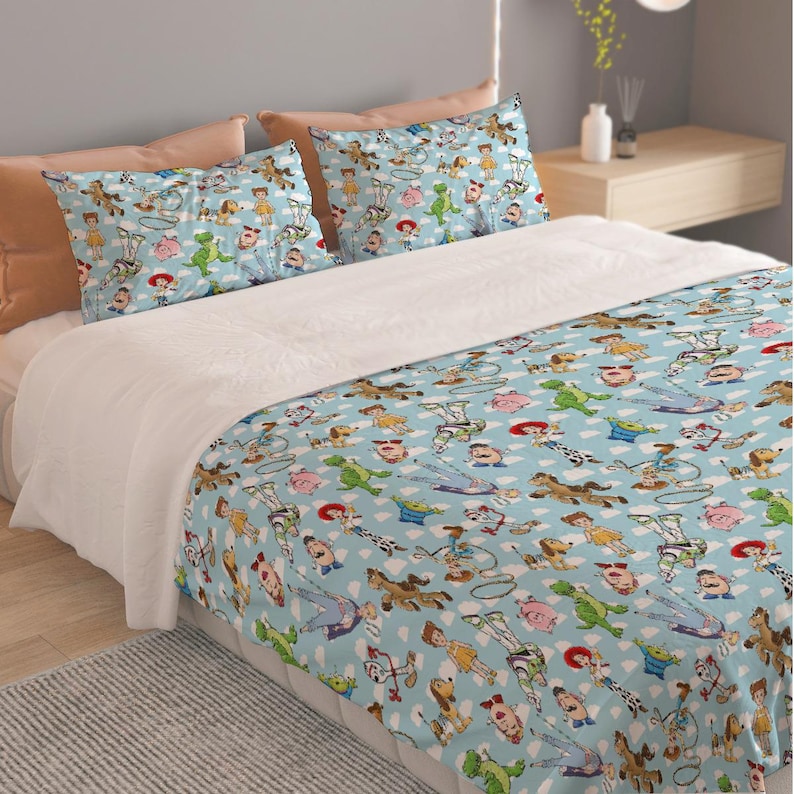 May include: A light blue bed set featuring a repeating pattern of cartoon characters, including a cowboy, a space ranger, and a green dinosaur. The set includes a comforter and two pillowcases.