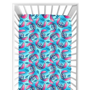 Peut inclure: Un drap de lit pour bébé avec un motif répété du personnage Stitch de Lilo & Stitch. Le motif comprend le visage et les oreilles de Stitch en rose et bleu, sur un fond bleu clair avec des éclairs roses.