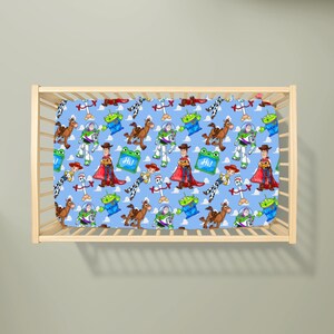 May include: A light blue cot sheet featuring a repeating pattern of Toy Story characters including Woody, Buzz Lightyear, Jessie, Bullseye, Forky, and green aliens, along with small green squares containing the text Hi in white lettering.