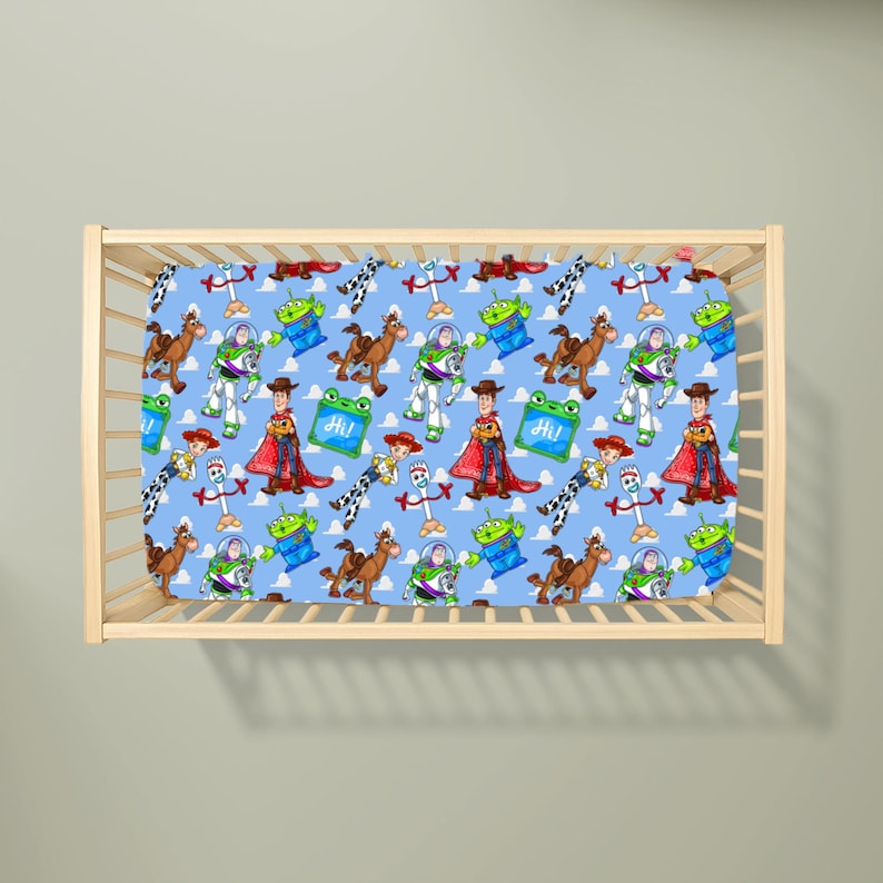 May include: A light blue cot sheet featuring a repeating pattern of Toy Story characters including Woody, Buzz Lightyear, Jessie, Bullseye, Forky, and green aliens, along with small green squares containing the text Hi in white lettering.