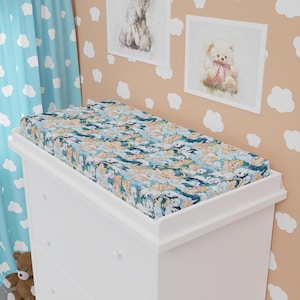 Könnte beinhalten: Ein weißer Wickeltisch mit einem Spannbettlaken mit einem Muster aus Teddybären in verschiedenen Farben. Das Laken liegt auf dem Wickeltisch. Der Hintergrund umfasst einen hellblauen Vorhang mit weißen Wolkenformen und gerahmte Teddybär-Kunst.