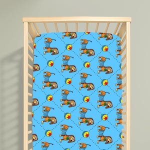 May include: A light blue cot sheet featuring a repeating pattern of brown dachshunds with blue spring bodies and colourful balls. The design is arranged in a diamond grid created by small dark blue dots, covering the entire surface of the fabric.