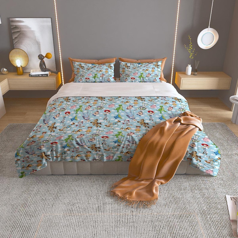 May include: A light blue comforter set featuring a repeating pattern of cartoon characters. The set includes a comforter, two pillow shams, and a brown throw blanket. The bedding is on a bed with a light gray base.