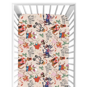 Peut inclure: Un drap-housse pour lit bébé avec un motif répété de personnages de dessins animés en tenue de cow-boy, de fers à cheval et les mots "Wild West" en beige. Le drap est dans un cadre de lit blanc.