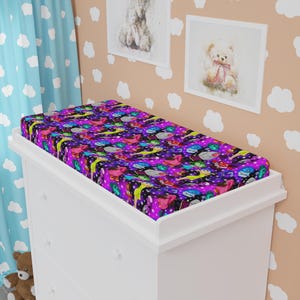 May include: A colorful changing pad cover with a vibrant space-themed pattern. The cover features a dark purple background with planets, rockets, and stars in various colors. It is placed on a white changing table, creating a playful nursery setting.