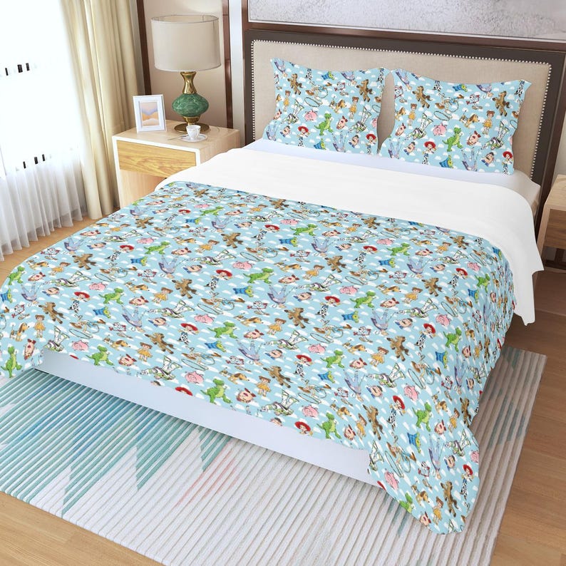 May include: A light blue bedspread and two pillowcases featuring a repeating pattern of cartoon characters. The bedding set has a white fitted sheet and is on a bed with a wooden headboard. The room has a neutral color scheme.