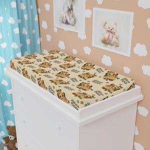 May include: A beige changing mat cover featuring a repeating pattern of a cartoon lion cub, stars, and leaves. The words "Hakuna Matata" are printed on the cover in a script font.
