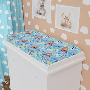 Könnte beinhalten: Eine Baby-Wickelauflage mit einem blauen Muster, das Cartoon-Figuren zeigt, darunter eine rothaarige Prinzessin, ein Schneemann und eine blonde Prinzessin. Die Auflage liegt auf einer weißen Kommode.