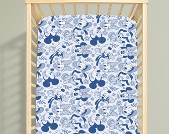 Puede incluir: Una sábana bajera de color azul claro con un patrón repetitivo de siluetas de Mickey Mouse en azul oscuro, caras de Daisy Duck y pequeños motivos florales. La tela está ajustada sobre una cuna de madera clara con barrotes verticales.