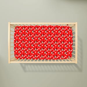 May include: A red fitted crib sheet with a repeating pattern of Minnie Mouse faces and white star shapes. The sheet is inside a light-colored wooden crib.