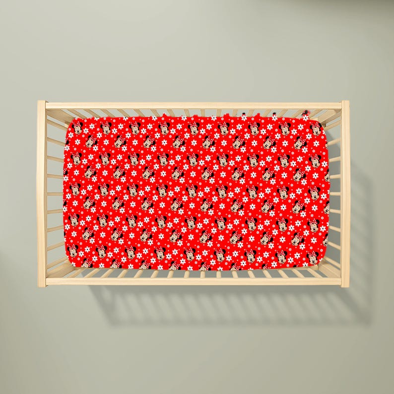 May include: A red fitted crib sheet with a repeating pattern of Minnie Mouse faces and white star shapes. The sheet is inside a light-colored wooden crib.