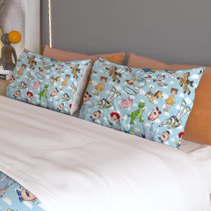 May include: Two light blue pillowcases with a repeating pattern of cartoon characters on a bed with white sheets. The pillowcases feature characters from the Toy Story franchise, including Woody, Buzz Lightyear, and Rex. The bed has orange pillows and a gray headboard.