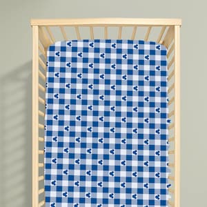 Könnte beinhalten: Ein Kinderbettlaken mit einem blau-weißen Gingham-Karomuster, das mit kleinen, dunkelblauen Mickey-Maus-Kopf-Silhouetten versehen ist, die gleichmäßig über den Stoff verteilt sind, gespannt auf einem hellen Holzgitterbett mit vertikalen Stäben.