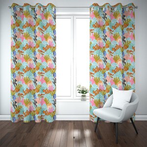 May include: A pair of patterned window curtains featuring a design of clownfish, coral, and other marine life in shades of orange, pink, and blue. The curtains are hanging in front of a window, with a white chair and a small plant visible.