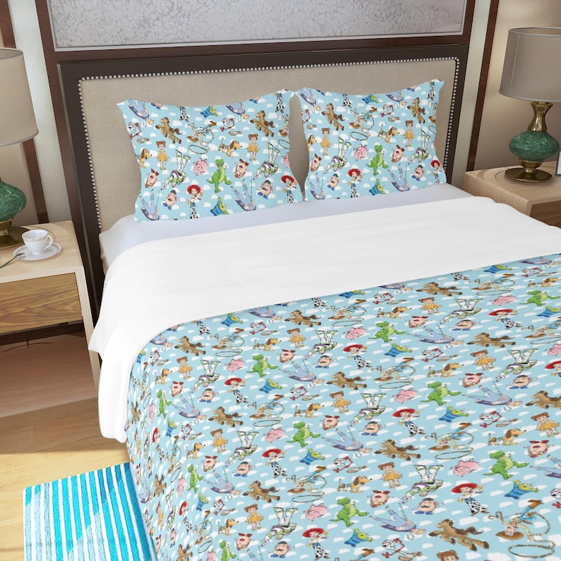 May include: A light blue bedding set featuring a repeating pattern of cartoon characters, clouds, and toys. The set includes a comforter and two pillowcases. The design is a whimsical and colorful illustration.
