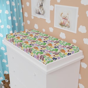 May include: A white changing table with a colorful changing pad. The pad features a repeating pattern of cartoon dinosaurs in shades of green, purple, orange, and brown on a cream background. The changing table is in a nursery setting.