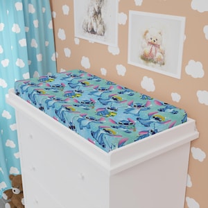 May include: A fitted changing pad cover featuring a repeating pattern of the character Stitch from the animated film Lilo & Stitch. The cover has a blue background with Stitch in various poses, accented with pink and yellow details.