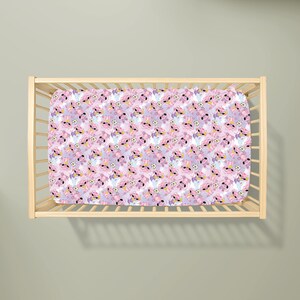May include: A crib sheet with a repeating pattern of a cartoon character in pink, purple, and white. The sheet is inside a wooden crib frame. The cartoon character has large eyes and a flower on its head. The background is a light purple color.