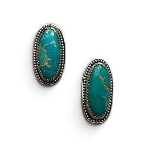 May include: A pair of oval-shaped stud earrings featuring polished turquoise stones with natural tan veining. Each stone is set in a silver bezel with a decorative beaded border, creating a textured metallic frame around the vibrant blue-green center.