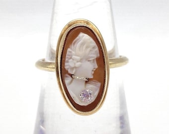 May include: Oval gold ring featuring a cameo of a woman's profile. The cameo is set in a brown background with a gold frame. A small, round, pink gemstone is set below the cameo. The ring is displayed on a clear stand.