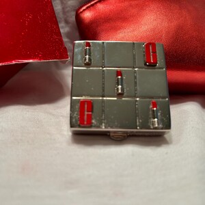 May include: A silver compact with a grid pattern and red lipstick designs. The compact is closed.