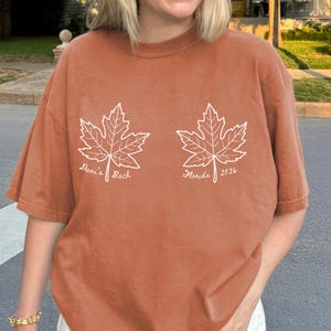 May include: A rust-colored short-sleeve t-shirt featuring two white line-art maple leaf graphics. The left leaf has the text 'Dani's Bach' and the right leaf has the text 'Florida 2026'. The shirt is paired with a white baseball cap and sunglasses.