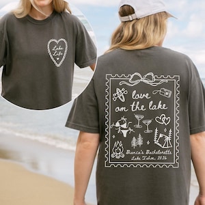 May include: Dark gray t-shirt with white graphic designs. The front features a heart with the words "Lake Life." The back has a framed design with the words "love on the lake" and other lake-themed illustrations. The text "Bianca's Bachelorette Lake Tahoe, 2026" is also included.