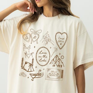 May include: Ivory-colored t-shirt with brown graphic illustrations, including hearts, a tent, a mountain, and a boat. Text includes "Love on the Lake," "Camp Alice," and "Alice's Bachelorette Lake Tahoe, 2026."