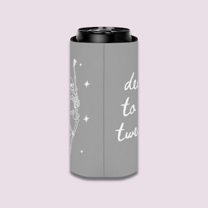 May include: A gray can cooler with a white skeleton graphic and the words "de to twe" in a script font. The skeleton is dancing with its arms raised. The background is a light pink.