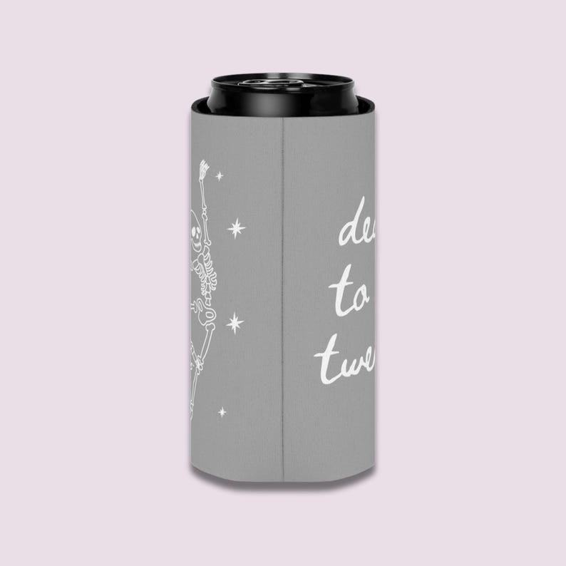 May include: A gray can cooler with a white skeleton graphic and the words "de to twe" in a script font. The skeleton is dancing with its arms raised. The background is a light pink.